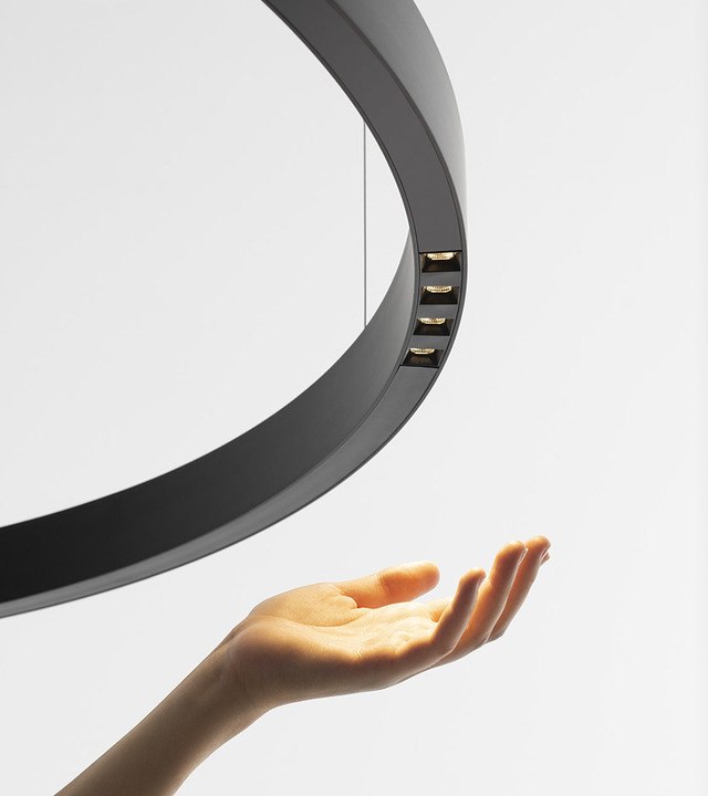 Lampada a sospensione circolare nera con moduli LED orientati verso il basso, con una mano che si alza sotto di essa.