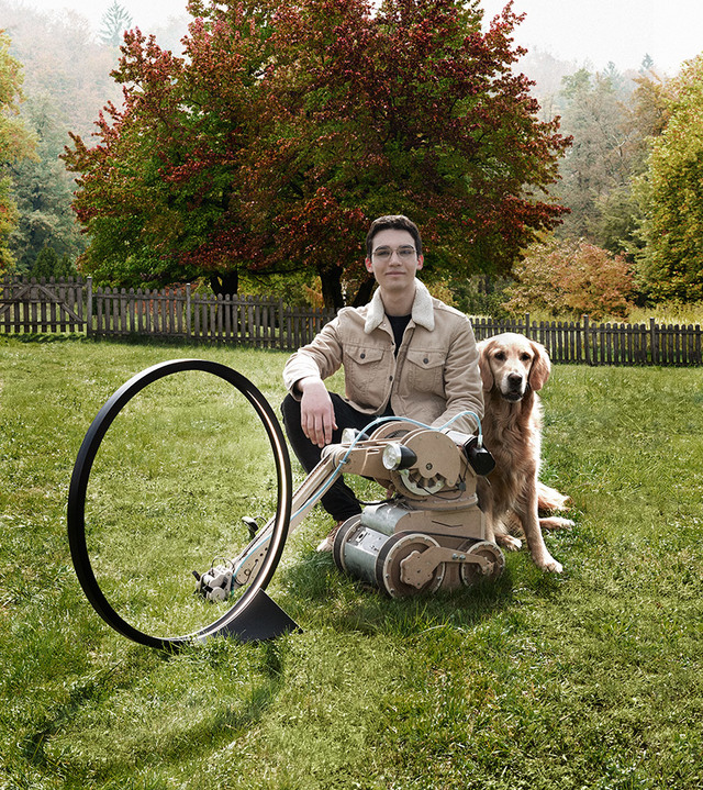 Giovane seduto sull'erba con un golden retriever accanto a una lampada da terra con paralume ad anello circolare, alberi autunnali sullo sfondo