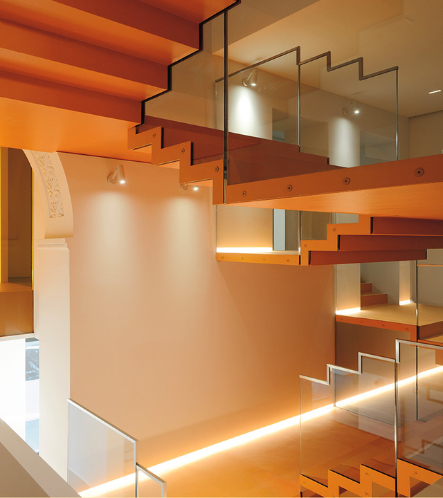 Modern staircase with orange wooden steps, glass railings, and warm Artemide LED strip lighting illuminating each tread from below.