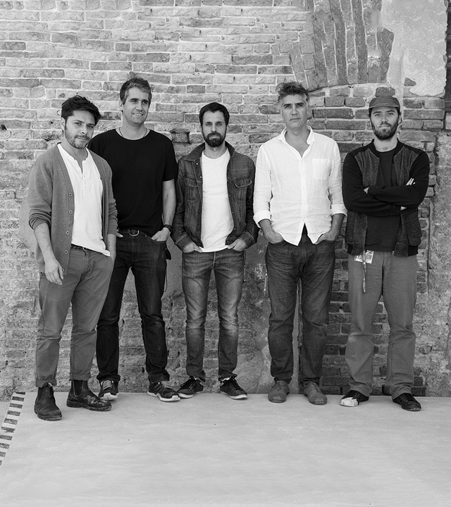 Cinque uomini in piedi insieme davanti a un muro di mattoni, in posa per un ritratto di gruppo in abbigliamento casual.