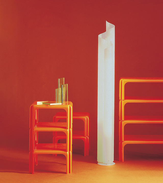 Tall white cylindrical floor lamp with organic curved top, displayed between orange modular furniture units against a red wall