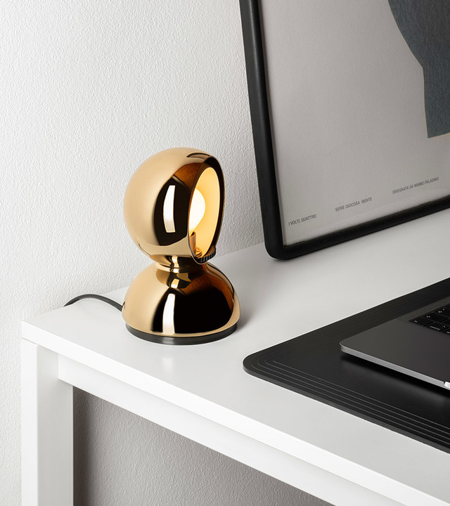 Artemide Eclisse table lamp in gold finish on a white desk, with reflective spherical shade revealing a warm glowing bulb.