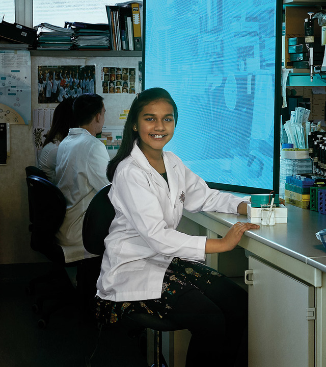 Ragazza giovane in camice da laboratorio sorridente a un banco di laboratorio, con un'altra studentessa che lavora sullo sfondo