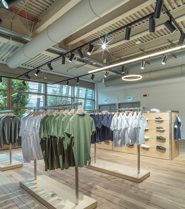 Interno di negozio di abbigliamento con faretti su binario e sospensione circolare Artemide che illuminano espositori in legno chiaro.