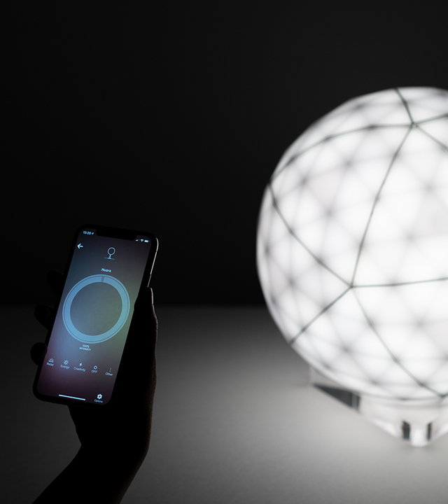Mano con smartphone e app Artemide che controlla una lampada da tavolo sferica luminosa con diffusore a griglia geometrica.