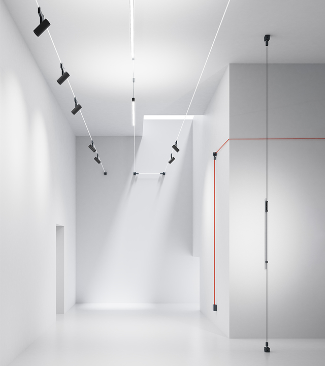 Sistema di illuminazione a cavi Artemide in uno spazio minimalista bianco: faretti neri regolabili su cavi tesi illuminano pareti e pavimento.