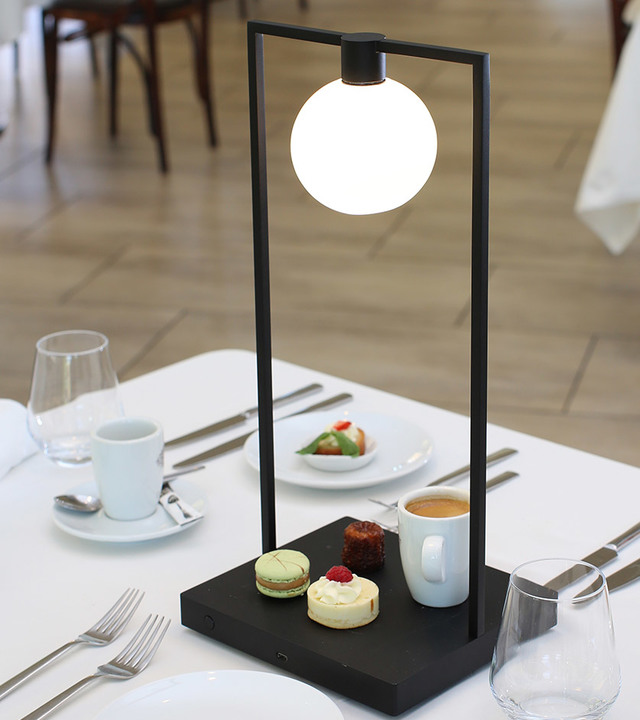 Lampada da tavolo Artemide nera rettangolare con diffusore globo opale su base scura, su tavolo da ristorante con pasticcini e caffè.