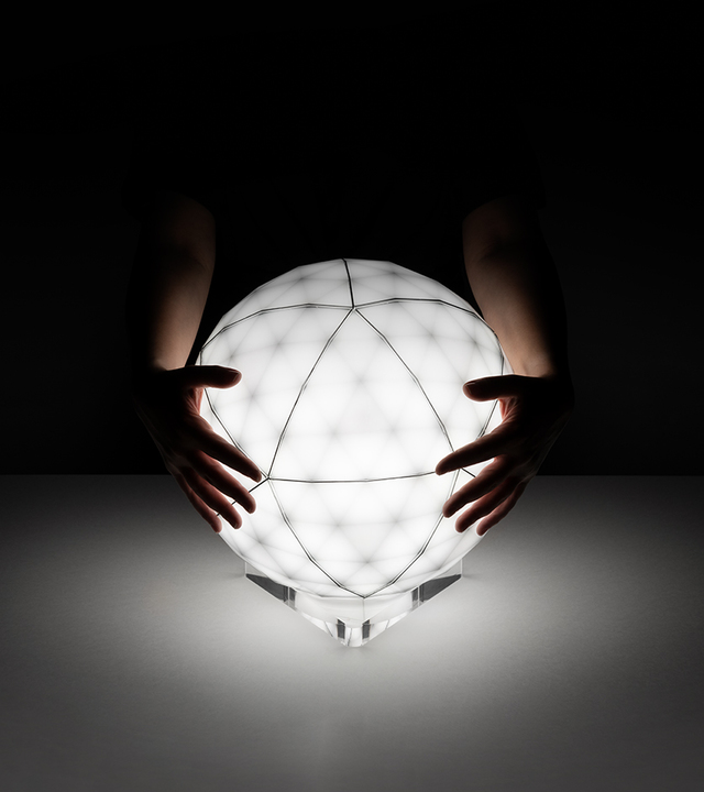 Mani che reggono una lampada sferica luminosa con superficie geometrica sfaccettata su base trasparente, che illumina una superficie scura.