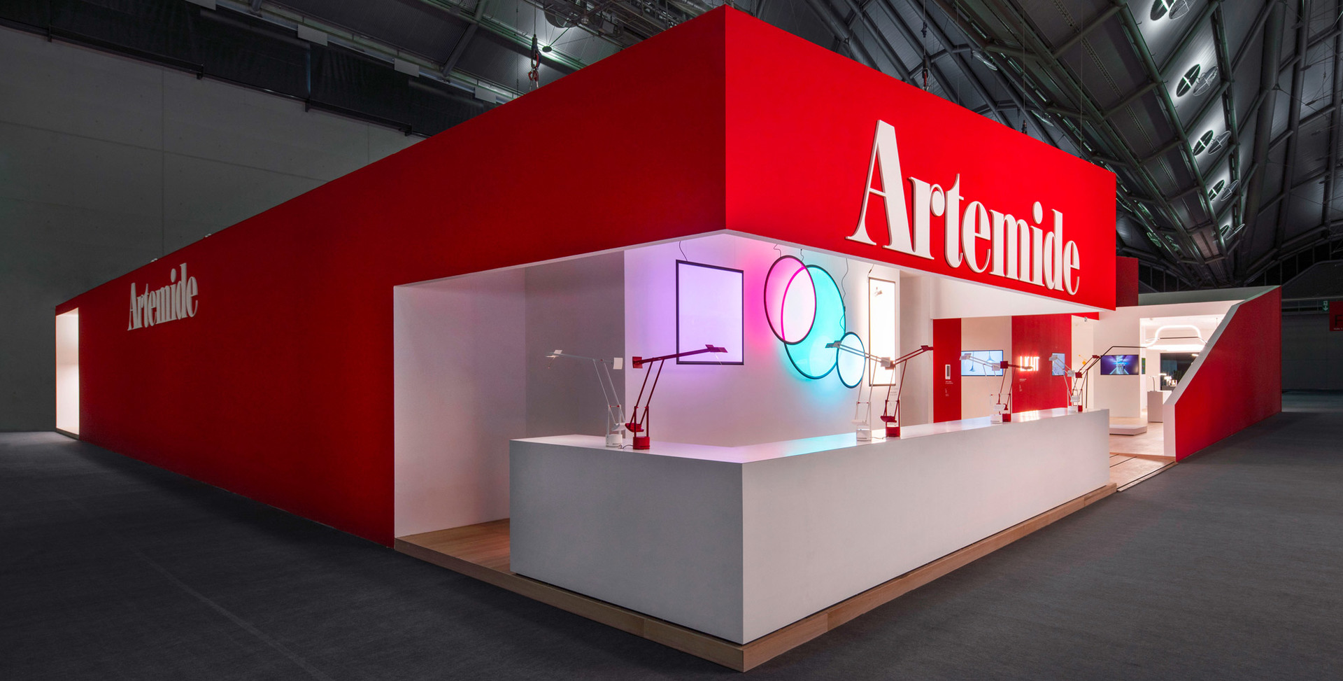 Artemide exhibition booth with bold red branding, white display counters, and colorful lighting installations showcasing the brand's lamp collection.
