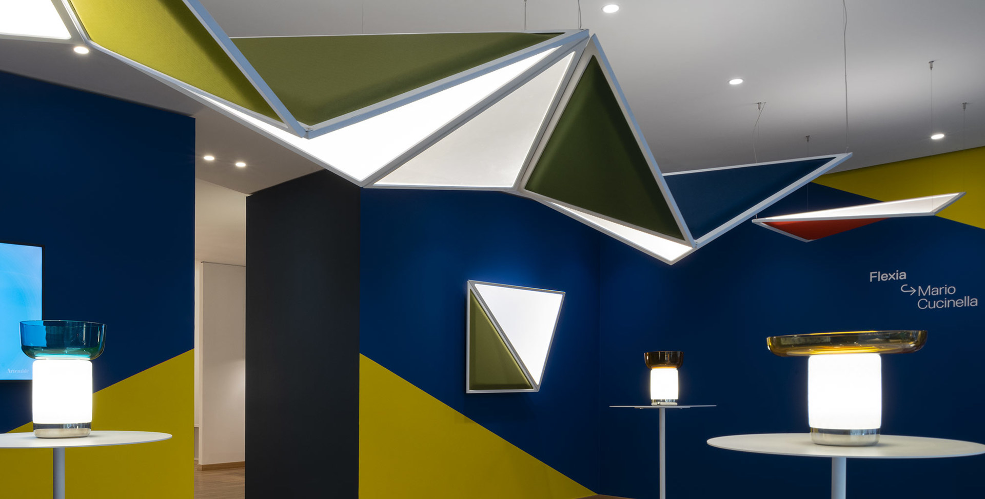 Artemide Flexia di Mario Cucinella: pannelli geometrici a soffitto e parete con illuminazione integrata, abbinati a lampade da tavolo in uno showroom