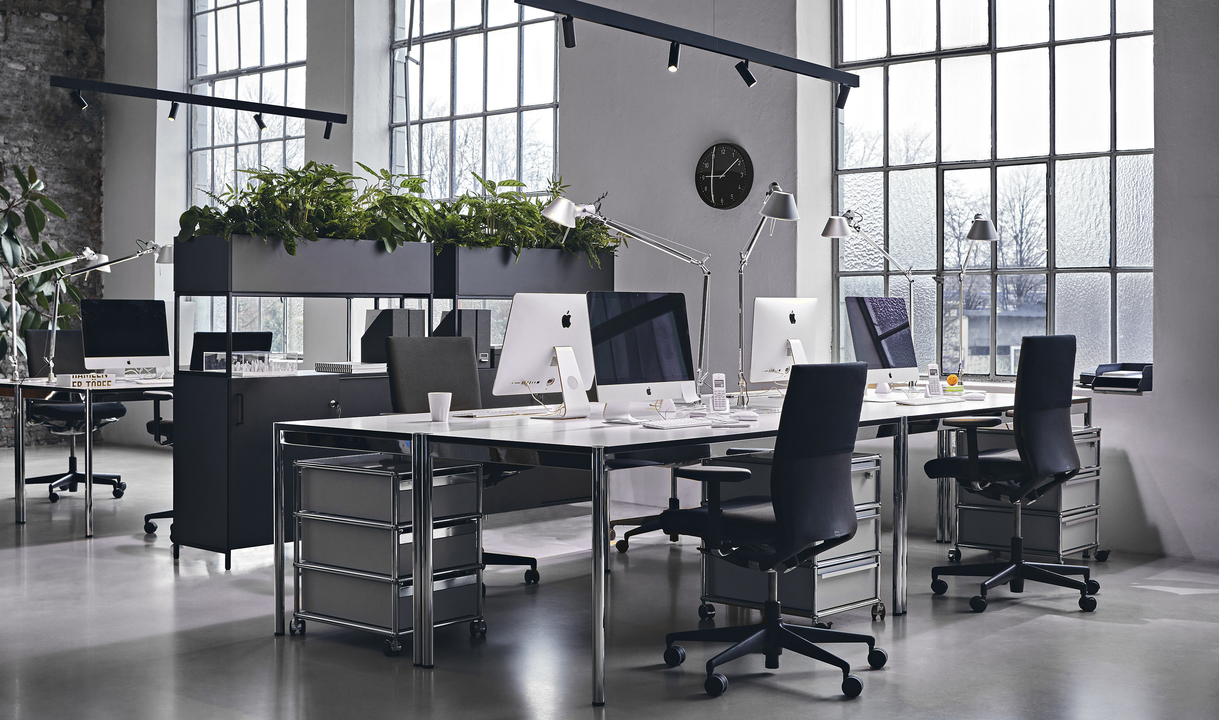 Ufficio open space moderno con lampade da tavolo Artemide Tolomeo, postazioni di lavoro, sedie nere, grandi finestre e divisori con piante verdi