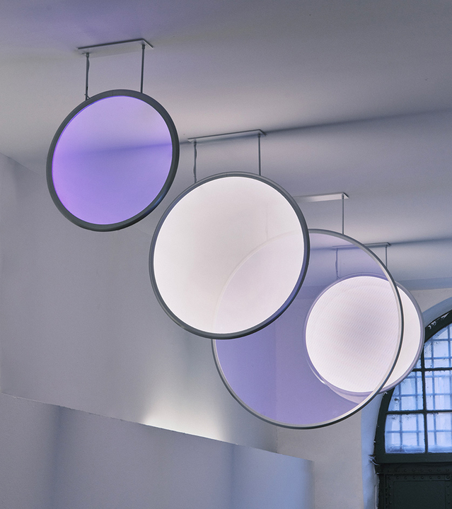 Three large circular pendant lights with dark frames, emitting white and purple light, suspended from ceiling mounts in a bright room.