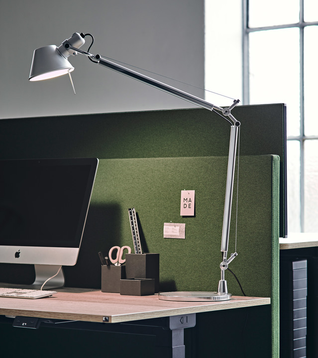 Lampada da tavolo Artemide Tolomeo in alluminio su una postazione di lavoro moderna, con braccio regolabile esteso sopra un monitor e pannello acustico verde.