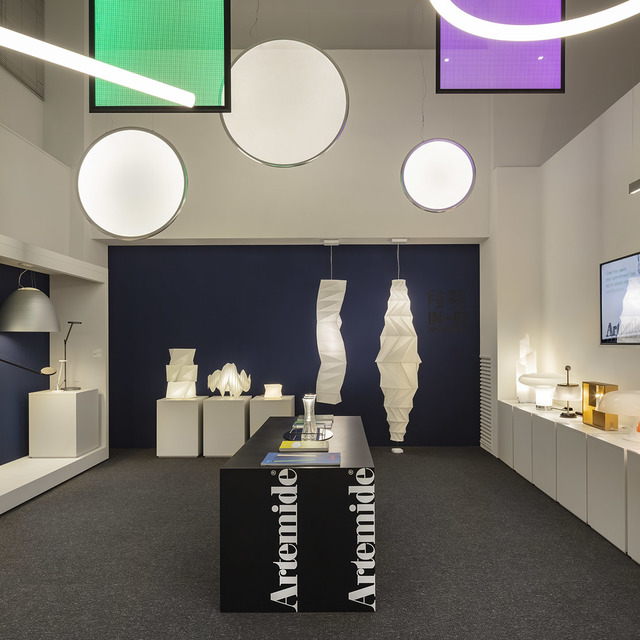 Artemide showroom displaying circular ceiling lights, sculptural white pendant lamps, and table lamps on white pedestals and shelving.