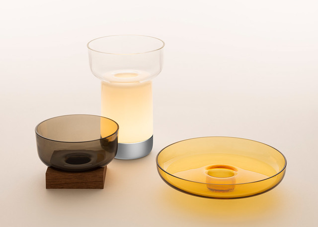 Collezione lampade da tavolo Artemide: ciotola in vetro fumé su base in legno, lampada cilindrica luminosa e piatto in vetro ambra con supporto centrale