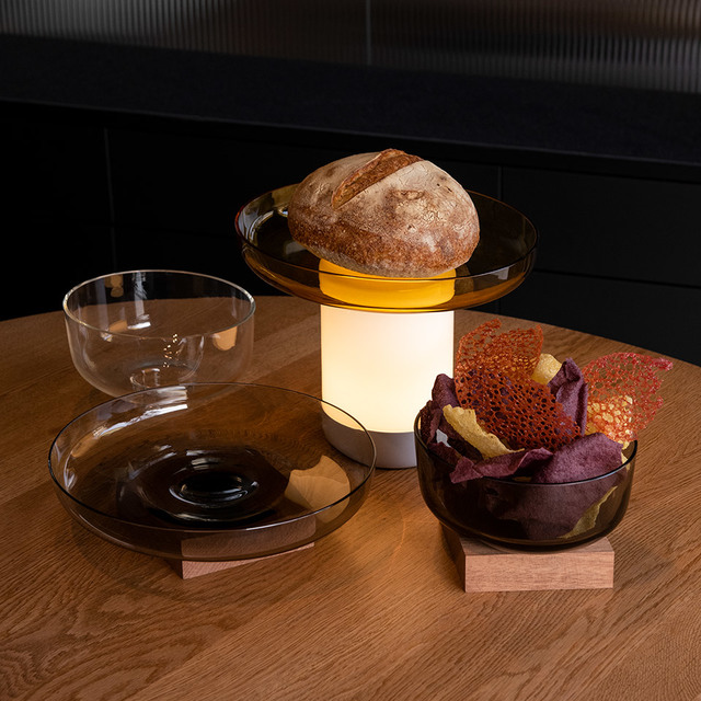 Lampada portatile Artemide Unplugged con luce bianca calda su tavolo in legno, circondata da ciotole di vetro con pane e snack