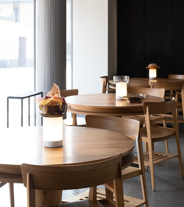 Sala da pranzo di ristorante con tavoli rotondi e sedie in legno, con lampade da tavolo portatili Artemide con paralumi cilindrici bianchi che diffondono luce calda.