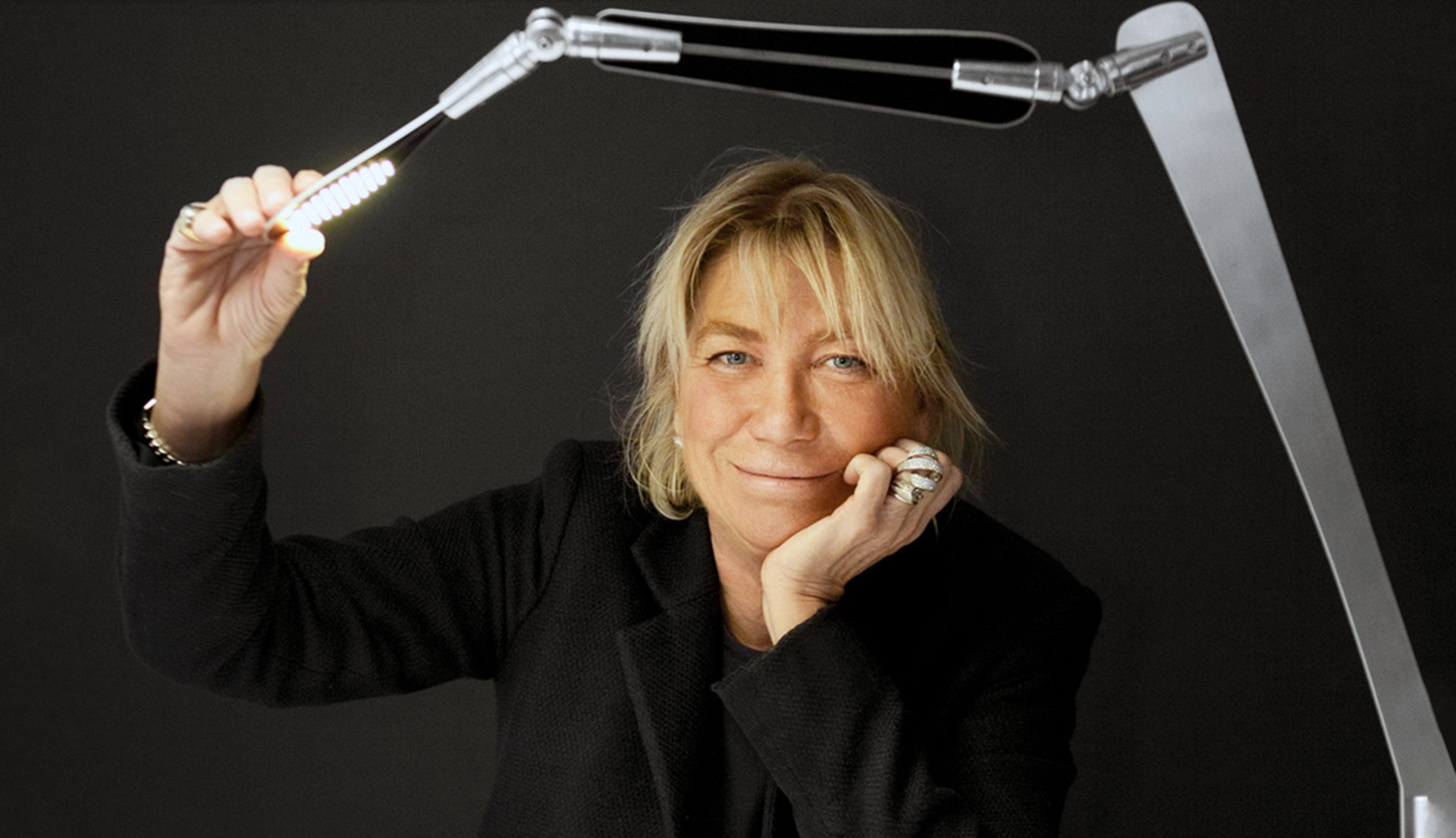 Woman posing with an Artemide adjustable LED desk lamp, holding its articulated arm while resting her chin on her hand.