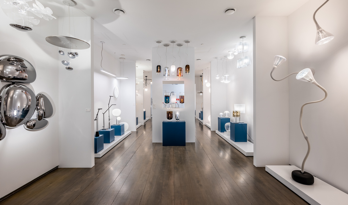 Showroom Artemide con pareti bianche, pavimento in legno scuro, lampade a sospensione, da tavolo e da terra su piedistalli blu
