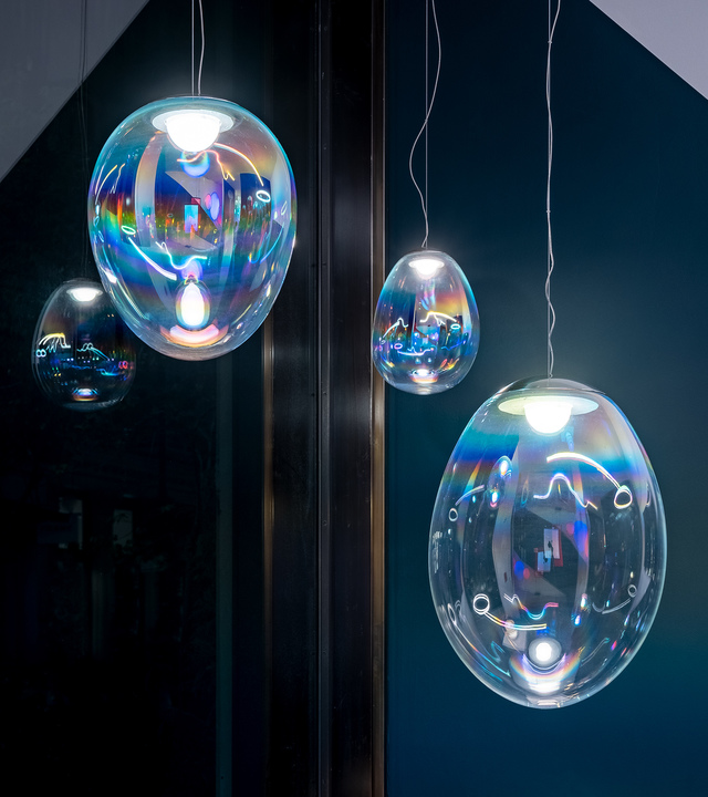 Quattro lampade a sospensione in vetro iridescente di diverse dimensioni ovali, che riflettono sfumature arcobaleno su sfondo verde petrolio scuro.