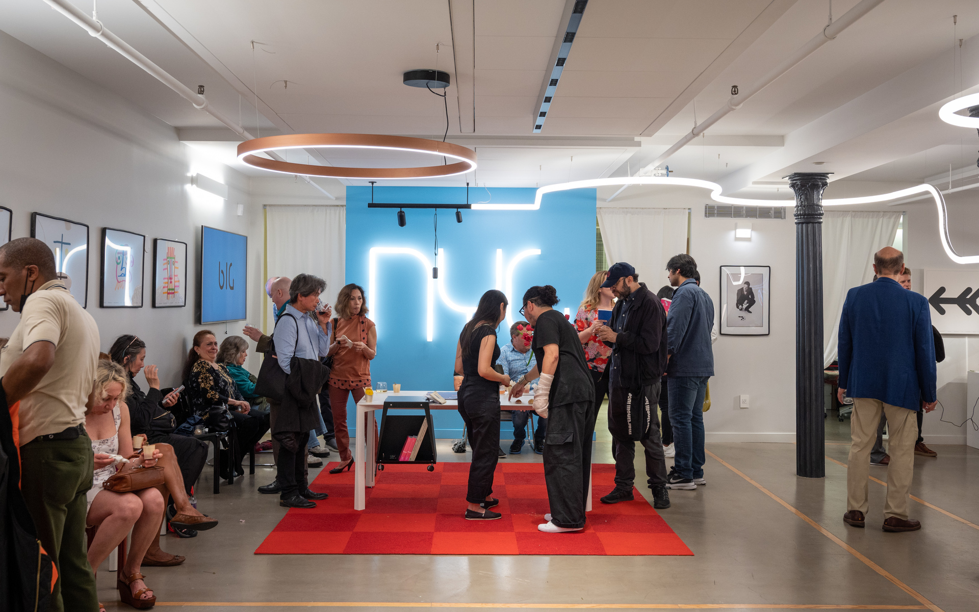 Evento in galleria con sospensione circolare Artemide e arte al neon; ospiti intorno a un tavolo su tappeto rosso sotto luci calde.