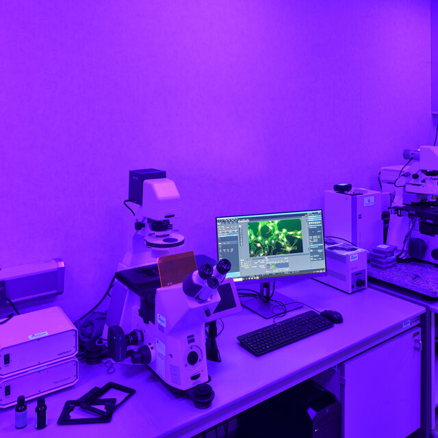Laboratory workstation bathed in purple light, featuring microscopes, a computer monitor displaying cell imagery, and scientific equipment on a white desk.