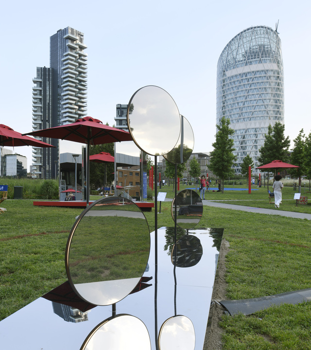 Sculture a specchio circolare lucido su steli metallici in un parco urbano di Milano, con grattacieli moderni e ombrelloni rossi sullo sfondo