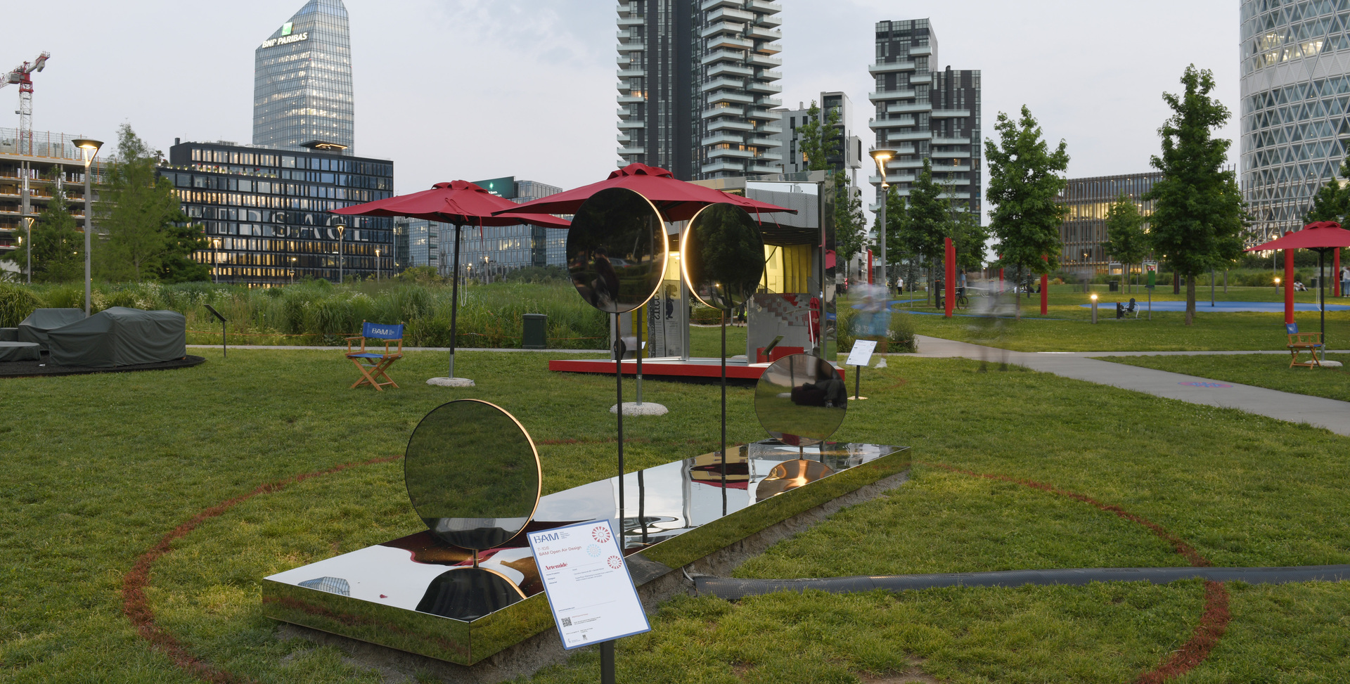 Installazione artistica esterna con specchi circolari e dettagli rossi su prato, con grattacieli moderni di Milano in un parco urbano.