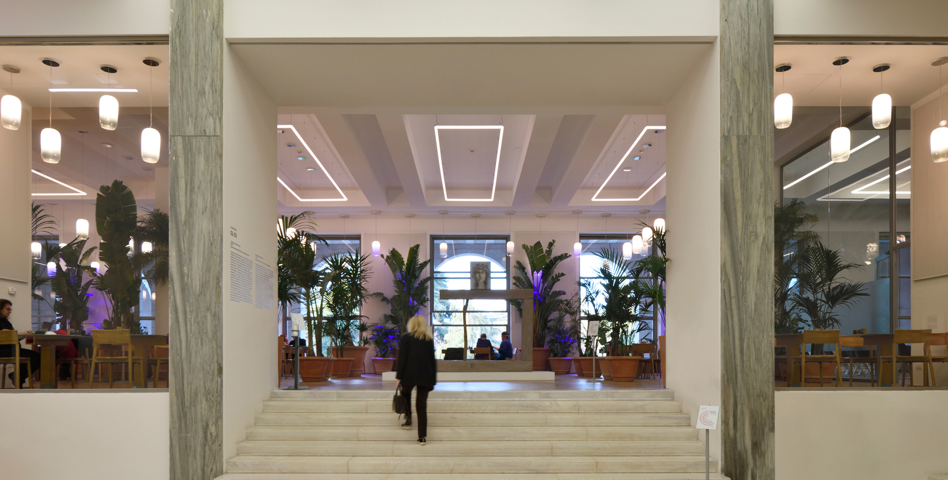 Ingresso di una grande hall con colonne in marmo, gradini e lampade a sospensione Artemide che illuminano lo spazio con luce calda ed elegante.