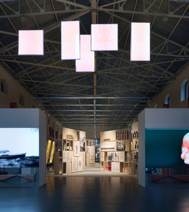 Sala espositiva con grandi pannelli luminosi rettangolari sospesi da soffitto a traliccio industriale, che illuminano i corridoi espositivi sottostanti