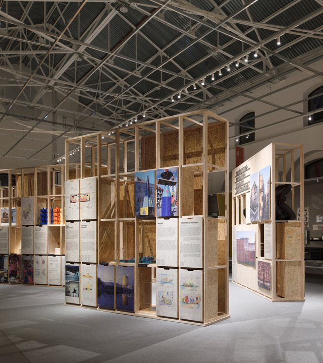 Spazio espositivo con unità modulari in legno che espongono pannelli architettonici colorati e tavole di progetto in un capannone industriale.
