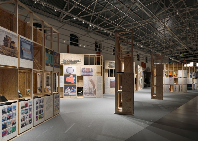 Sala espositiva con strutture modulari in legno che mostrano poster e oggetti, illuminata da faretti su binario a soffitto in uno spazio industriale