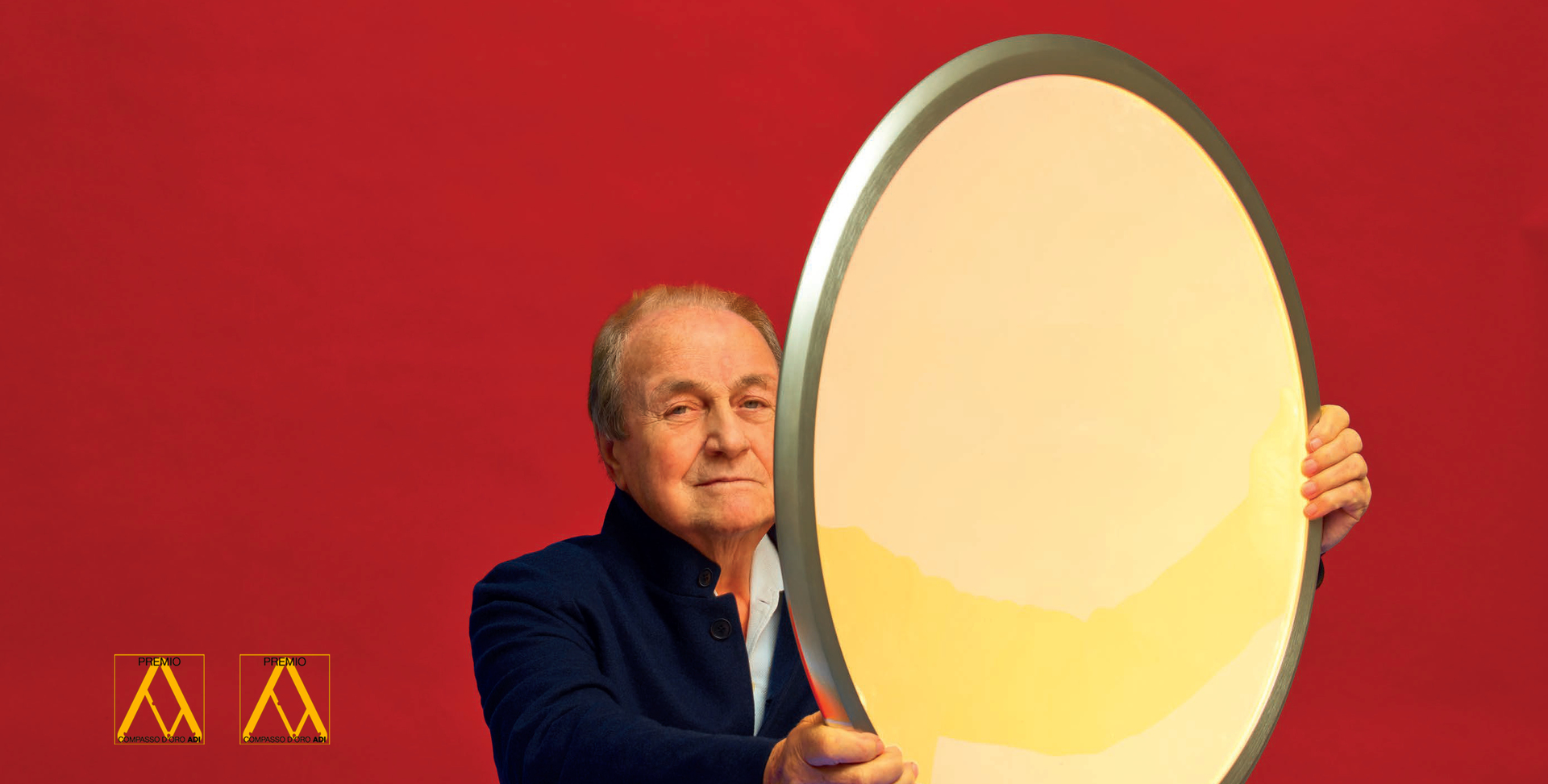 Designer che regge una grande lampada Artemide ovale con diffusore bianco e cornice argento su sfondo rosso.
