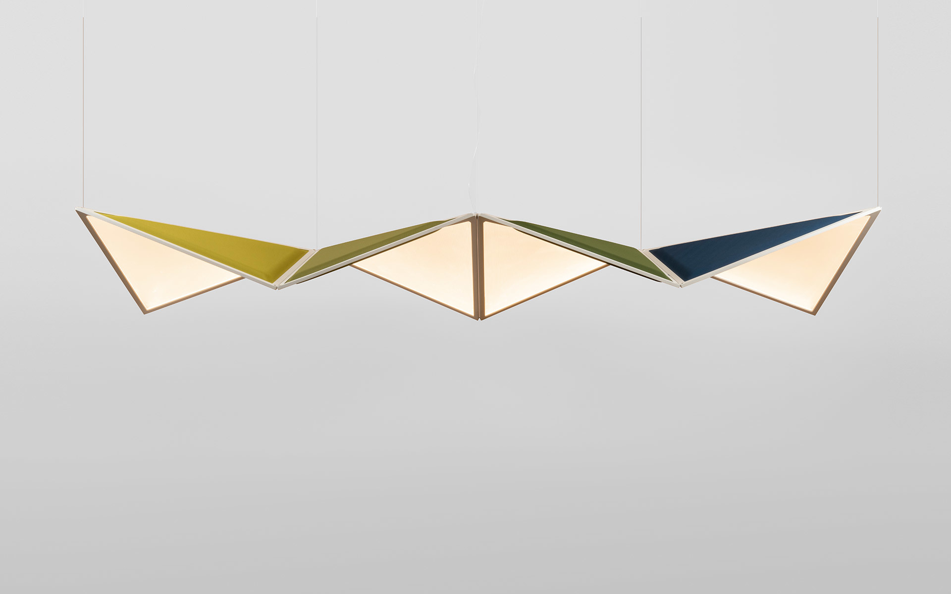 Tre lampade a sospensione geometriche a forma di rombo in giallo, verde oliva e blu navy, con diffusori angolari che emettono luce calda