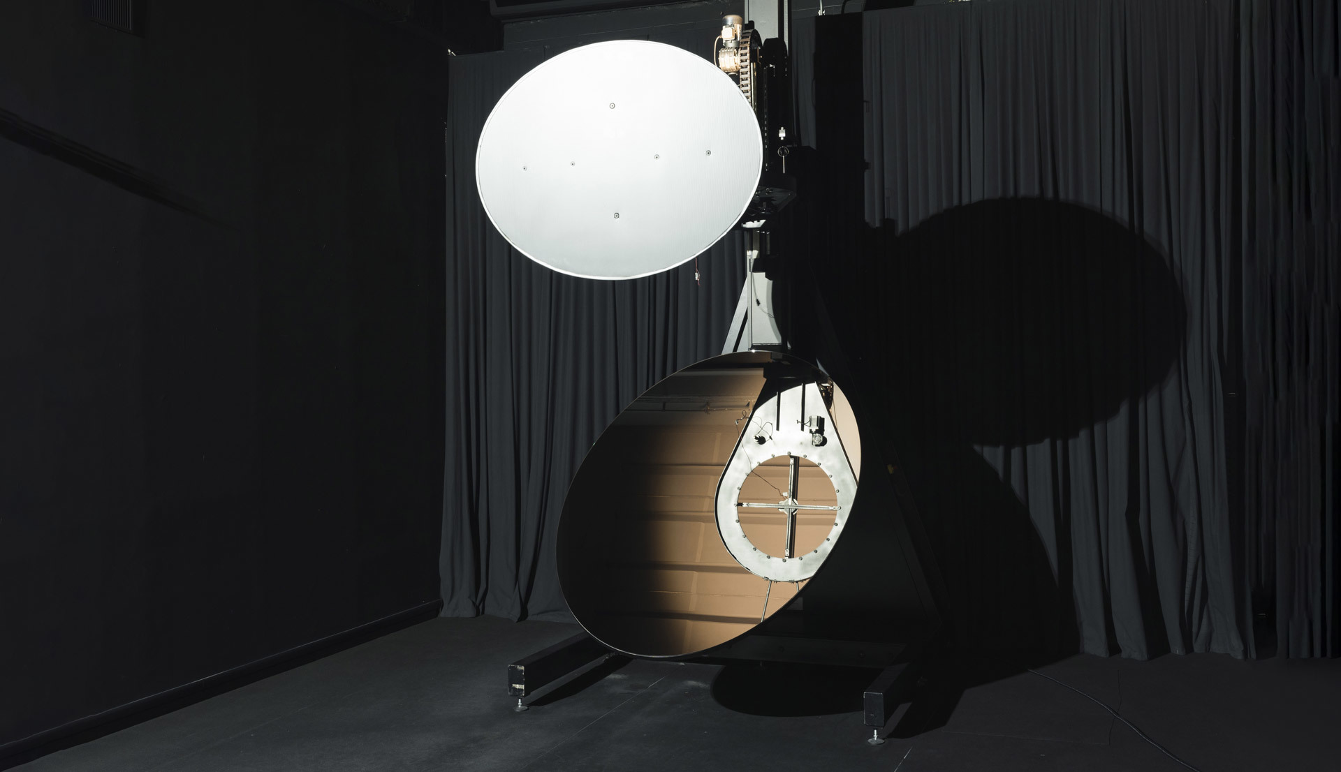 Prototipo di lampada Artemide con grande diffusore circolare bianco montato su supporto accanto a una struttura ovale in legno in uno studio buio