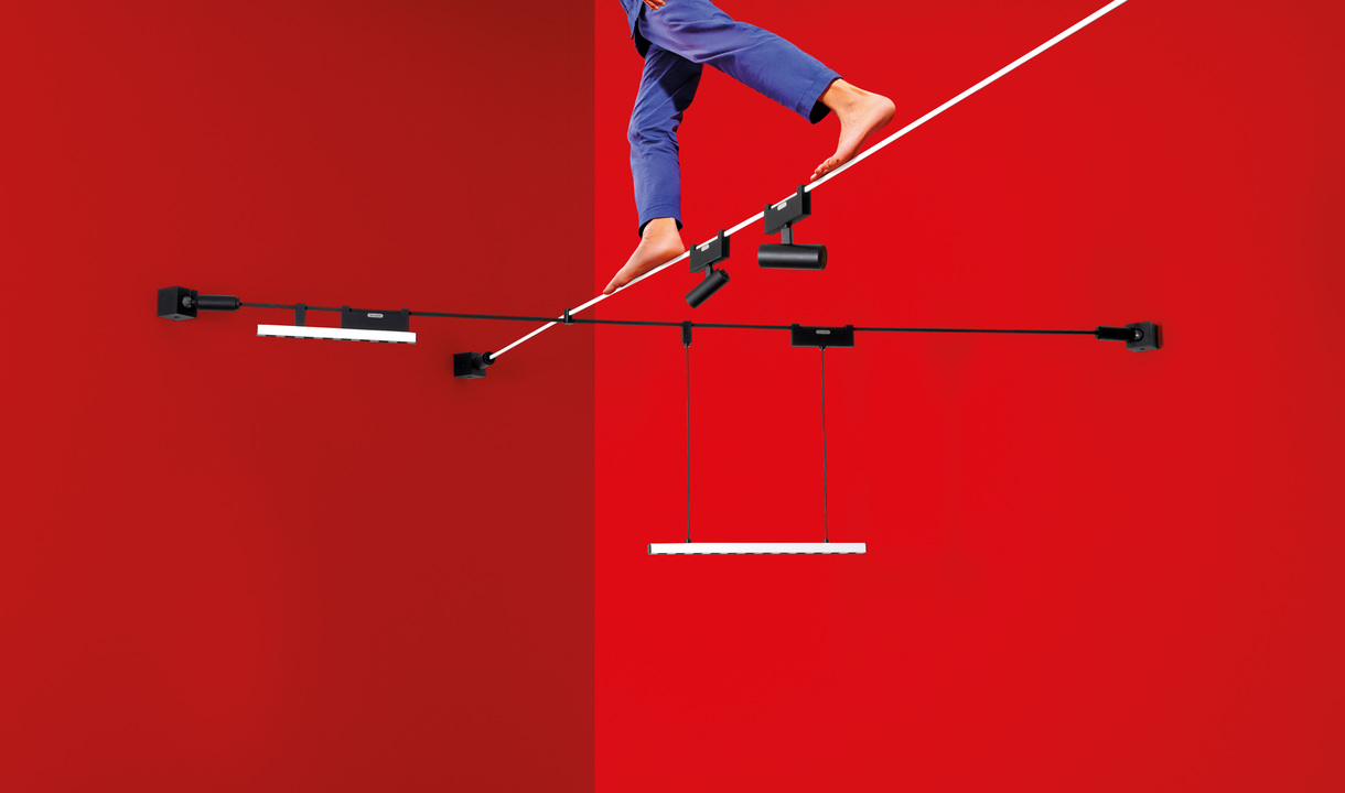 Artemide cable lighting system with black track, pendant and wall-mounted LED fixtures against a red background, with a person balancing above