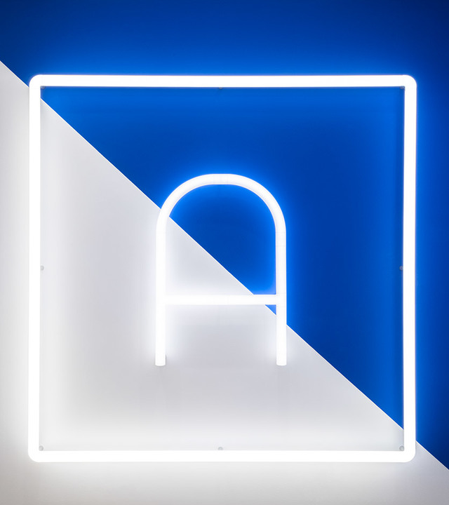 Logo Artemide in luce al neon bianca a forma di lettera A con arco, incorniciato da quadrato luminoso, su sfondo diagonale blu e bianco
