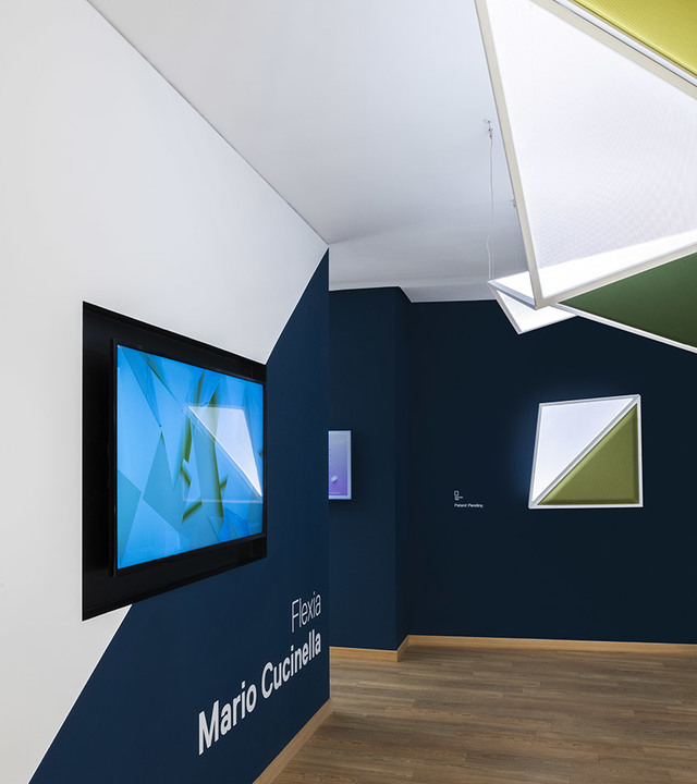 Lampada Artemide Flexia di Mario Cucinella esposta in uno spazio espositivo blu scuro con installazione geometrica a soffitto.