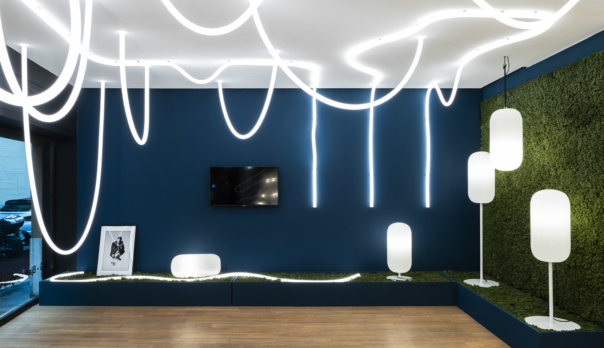 Showroom Artemide con luci a soffitto LED curvilinee, lampade da terra e a sospensione ovali bianche, su parete blu scuro e pannello di muschio verde.