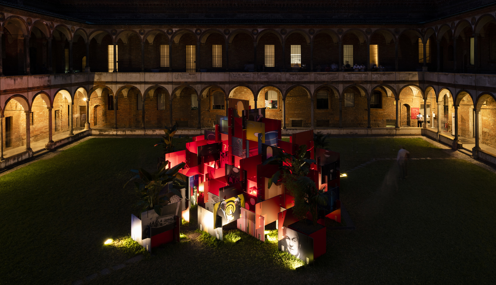 Illuminated red cubic installation in a historic courtyard at night, with Artemide ground lights highlighting surrounding greenery and archways.
