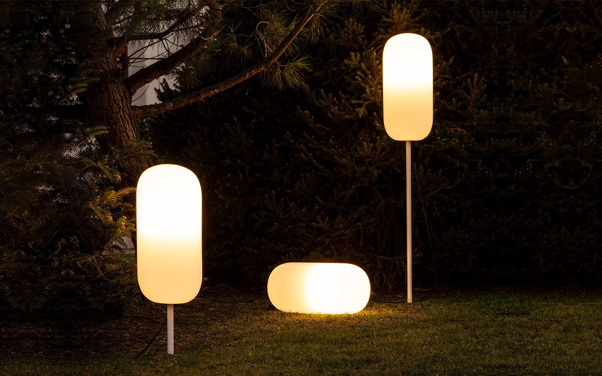 Tre lampade da esterno Artemide con diffusori bianchi luminosi su un prato al crepuscolo: due alte su palo e una bassa a terra.