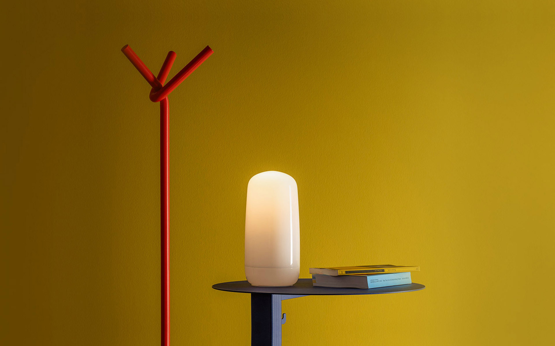 Lampada portatile ovale bianca luminosa su un tavolino scuro, accanto a libri, su sfondo giallo senape con appendiabiti rosso.