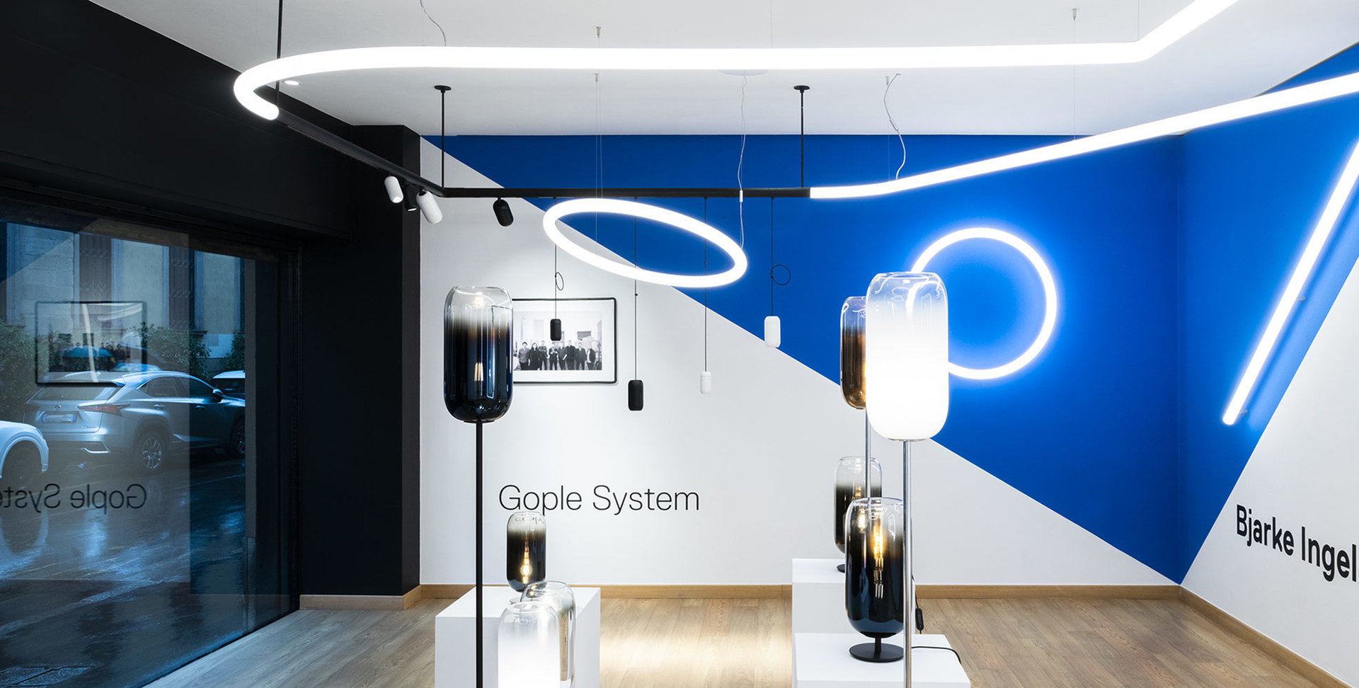 Showroom Artemide con lampade da terra Gople System con paralumi in vetro fumé e luci a sospensione circolari su parete blu e bianca.