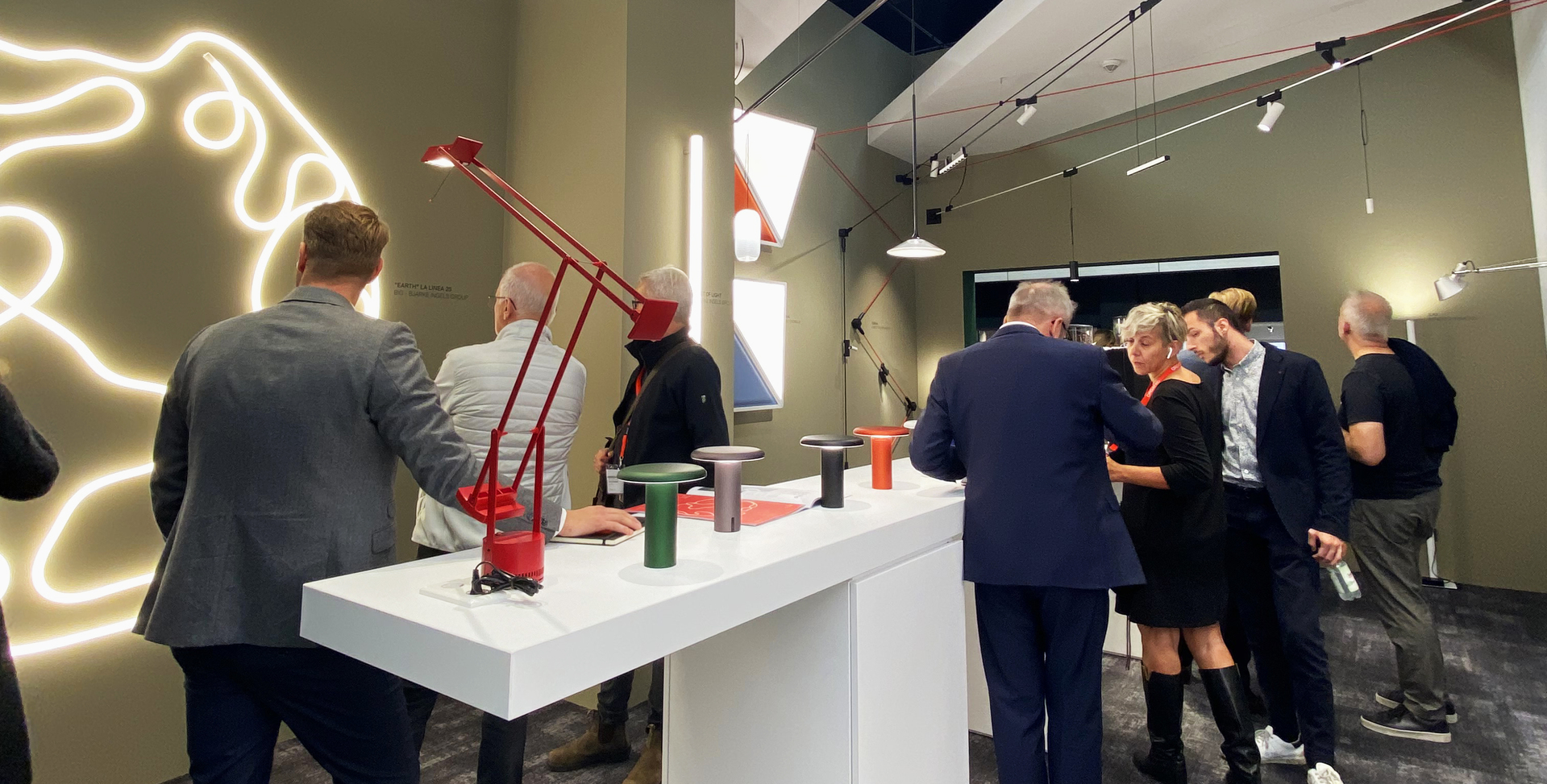 Visitors exploring Artemide lighting collection at a showroom, featuring a red desk lamp and small table lamps displayed on a white counter.