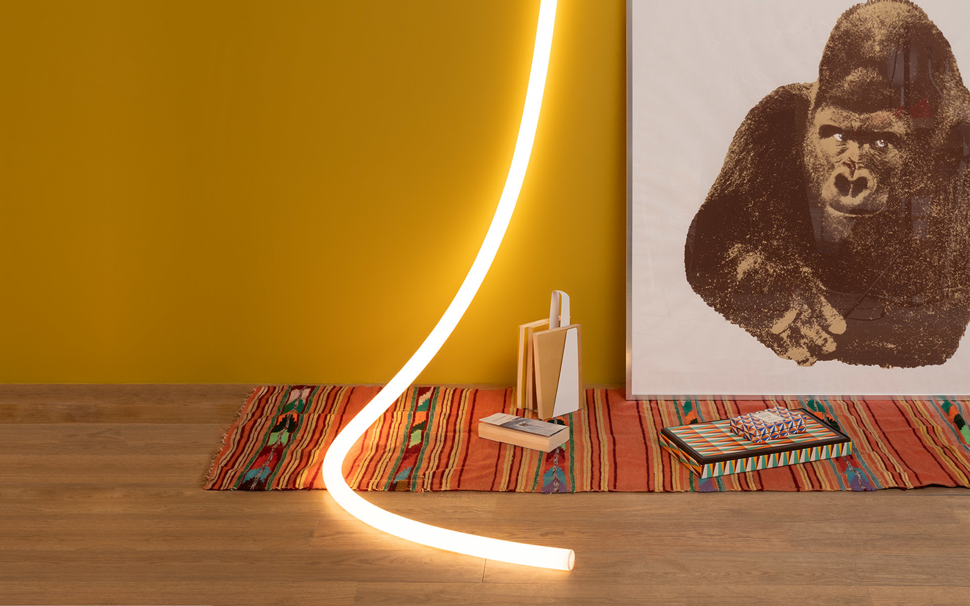 Tubo luminoso LED flessibile Artemide che si curva dal pavimento alla parete, luce bianco caldo, accanto a tappeto colorato e opera con gorilla