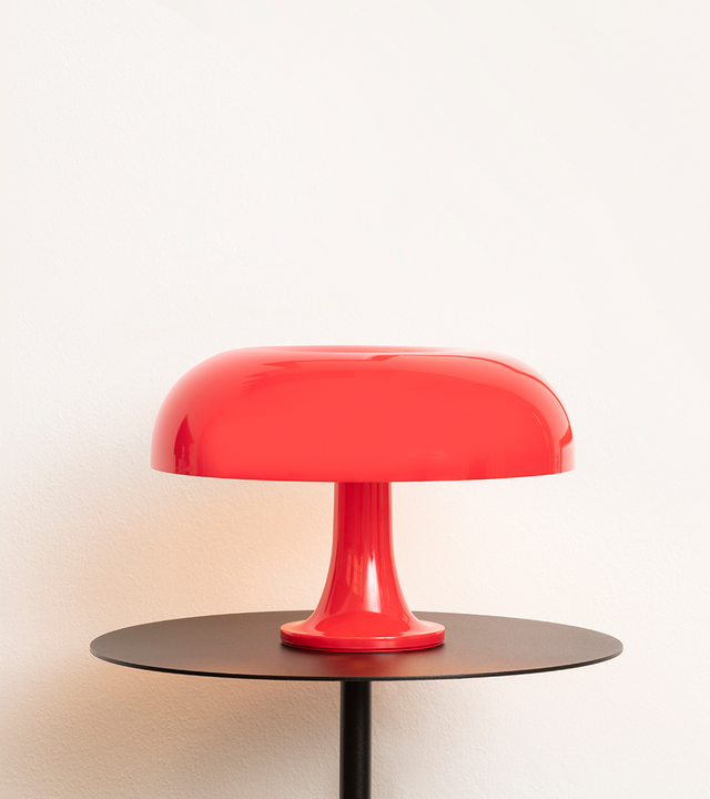 Lampada da tavolo Artemide Nessino in rosso, con iconico paralume a fungo su base a piedistallo, posata su un tavolo rotondo scuro