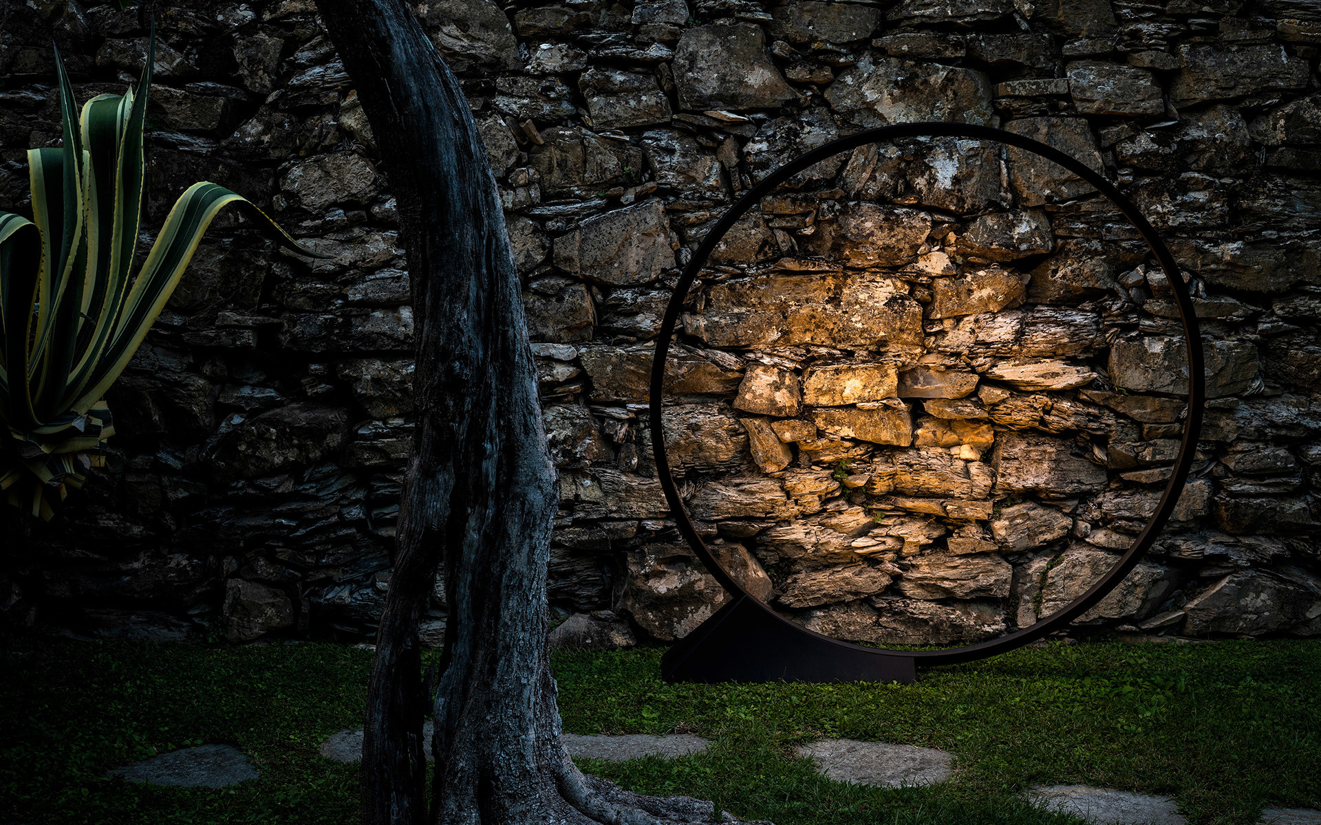 Lampada da terra in metallo nero circolare che illumina un muro in pietra rustica in un giardino esterno di notte, con pianta di agave vicina