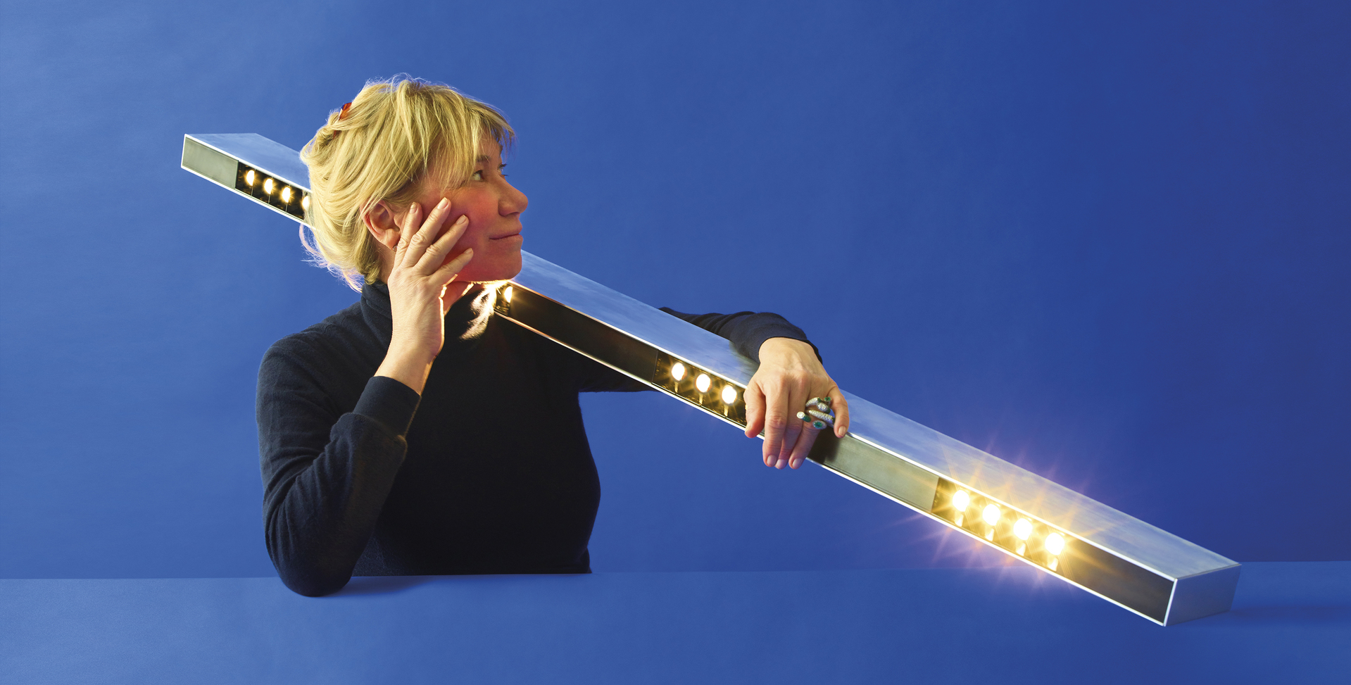 Donna che regge una lunga barra LED rettangolare con LED bianco caldo accesi, che mostra il design dell'illuminazione lineare Artemide.