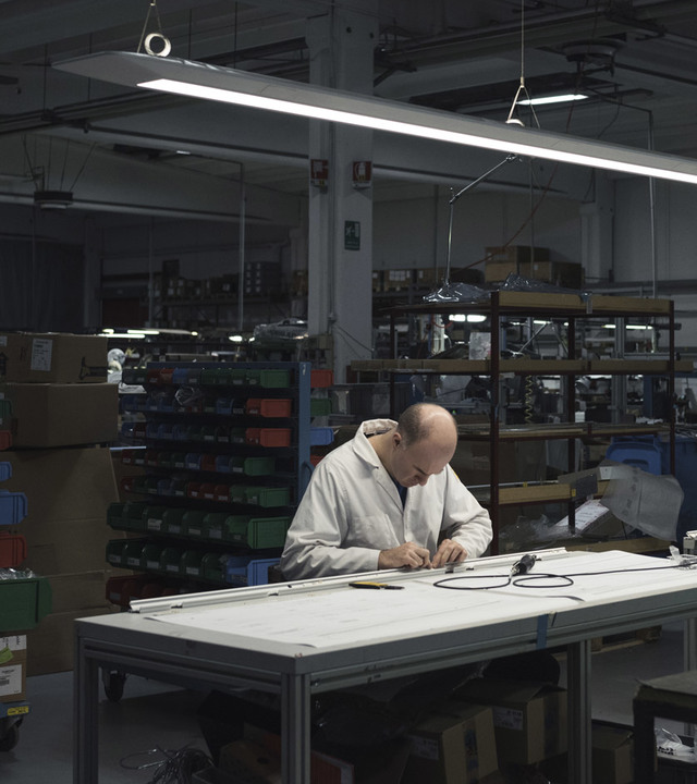 Tecnico che assembla un apparecchio illuminante a un banco di lavoro nella fabbrica Artemide, illuminato da una lampada a sospensione LED lineare.