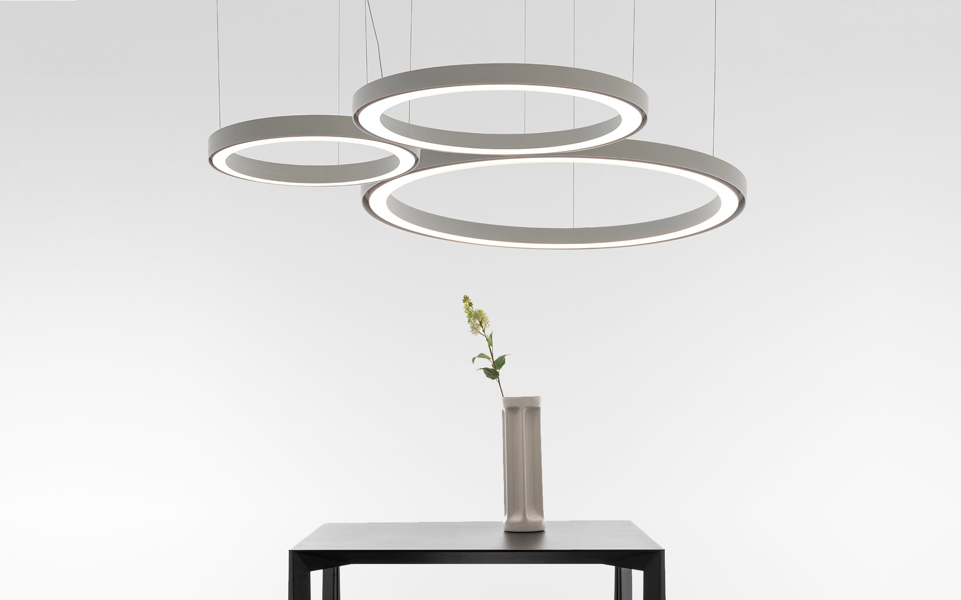Tre lampade a sospensione circolari LED di diverse dimensioni in grigio, sospese sopra un tavolo scuro con un vaso e un fiore.